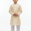 Young boy wearing beige kurta with white churidar, standing barefoot on white backdrop