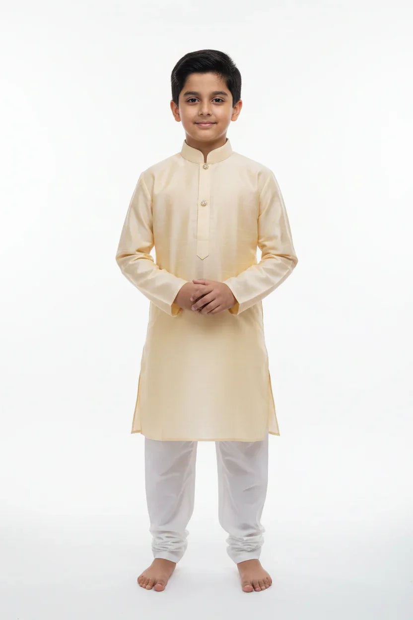 Young boy wearing beige kurta with white churidar, standing barefoot on white backdrop