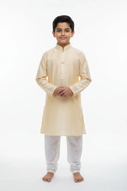 Young boy wearing beige kurta with white churidar, standing barefoot on white backdrop
