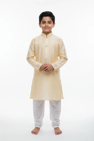 Young boy wearing beige kurta with white churidar, standing barefoot on white backdrop