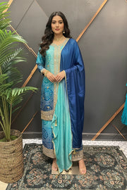Woman wearing aqua and blue embellished Indian ethnic suit with matching dupatta, posing indoors on floral rug