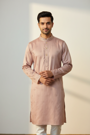 A man wearing a light pink long-sleeve kurta with a mandarin collar and white trousers, standing against a neutral light gray background.