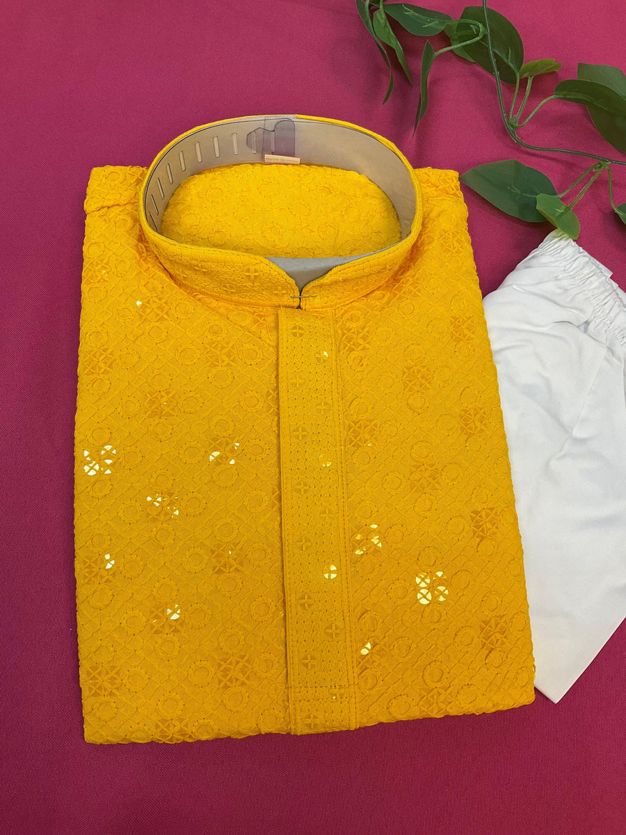 Folded yellow Indian ethnic wear kurta with sequin embroidery and white churidar on pink background with green leaves
