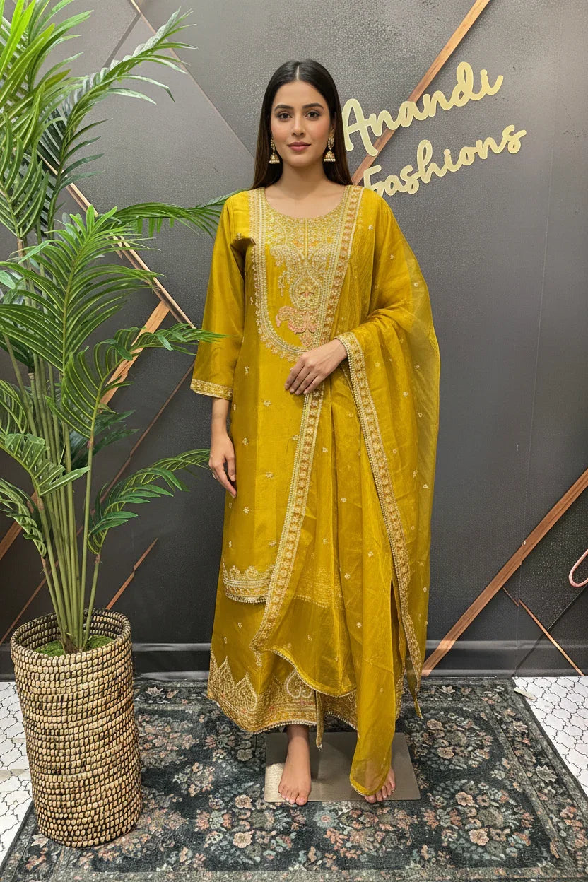 Woman wearing mustard yellow embroidered Indian ethnic suit with matching dupatta, standing barefoot on floral rug by a potted plant
