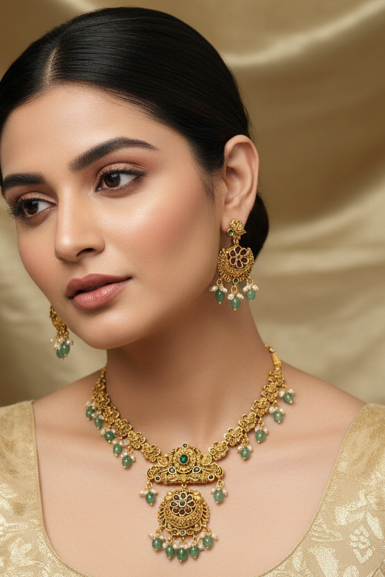 Close-up of woman wearing traditional gold Indian jewelry set with green beads, including necklace and earrings, against beige fabric background