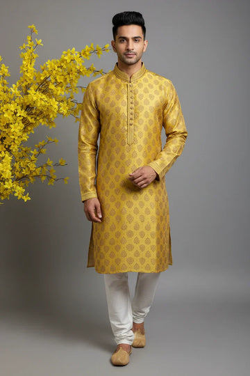 Man wearing golden yellow traditional Indian kurta with white churidar and jutti, standing by yellow flowers