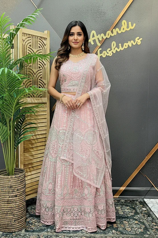 Woman in a pink traditional outfit standing in front of a decorative wall with 'Anandi Fashions' branding.