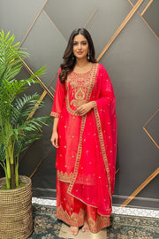 Woman in red embroidered Indian ethnic wear with gold detailing, standing indoors near a decorative wall and potted plant