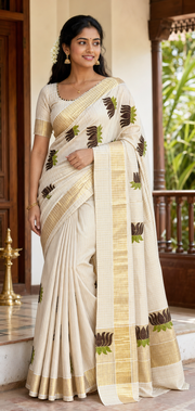 A woman modeling a cream-colored cotton saree with gold zari borders and brown and green floral motifs, standing in a traditional indoor setting with wooden railings.