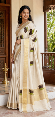 A woman wearing a cream-colored saree with gold zari borders and brown and green floral motifs, standing in a traditional indoor setting with wooden railings and a lamp.
