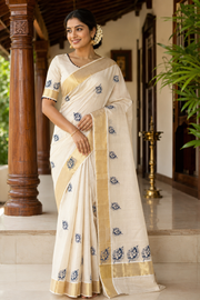 A woman wearing a cream-colored cotton saree with blue floral embroidery and a gold border, standing on a tiled porch with wooden pillars.