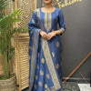 Woman wearing blue ethnic Indian salwar kameez with gold motifs and dupatta, standing next to wooden screen and plant at Anandi Fashions