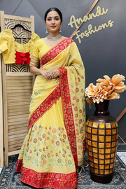 Woman in a yellow and red saree standing next to a decorative vase with flowers, against a branded backdrop.