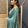 Woman in teal Indian saree with gold embroidery and border, styled hair, and gold earrings indoors