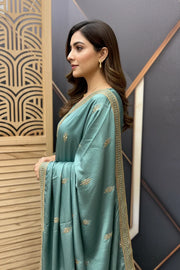 Woman in teal Indian saree with gold embroidery and border, styled hair, and gold earrings indoors