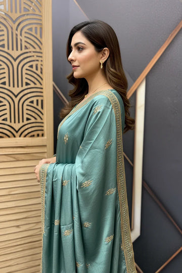 Woman in teal Indian saree with gold embroidery and border, styled hair, and gold earrings indoors