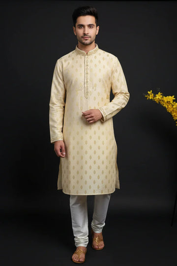 Man wearing beige embroidered kurta with white churidar and traditional sandals against black background