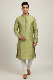 Man wearing green patterned Indian kurta with white churidar and brown sandals on plain background
