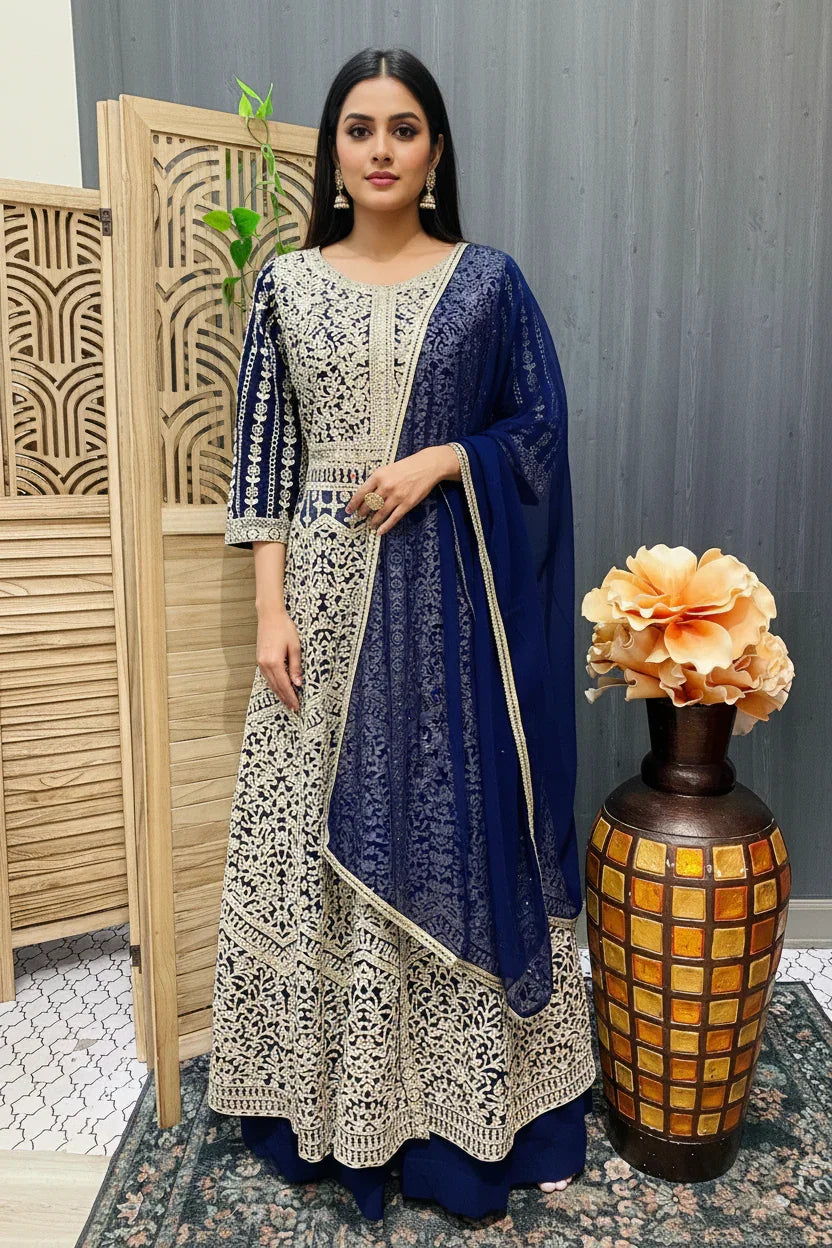 Woman in navy blue and beige embroidered Indian ethnic Anarkali dress with dupatta, standing indoors near wooden screen and decorative vase