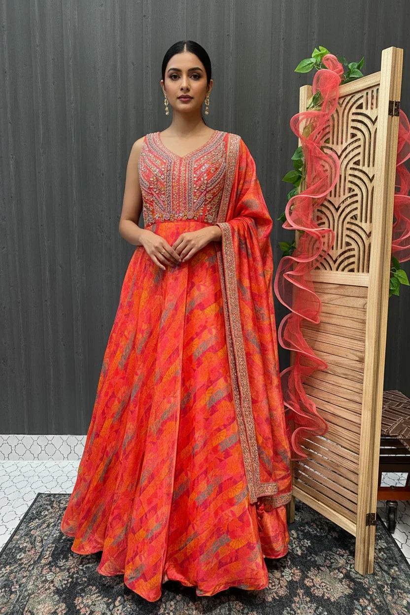 Woman wearing vibrant orange-pink embroidered Indian ethnic gown with matching dupatta, standing indoors