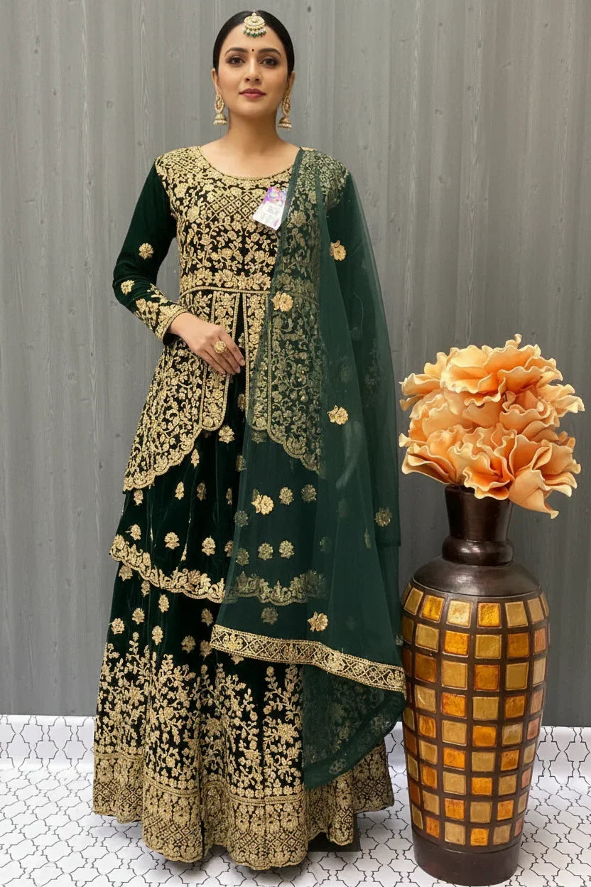 Woman wearing dark green and gold embroidered Indian ethnic wear with matching dupatta and gold jewelry, standing beside decorative vase