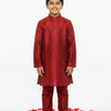 Smiling boy wearing red silk kurta pajama and beige traditional shoes, standing on white floor with red rose petals