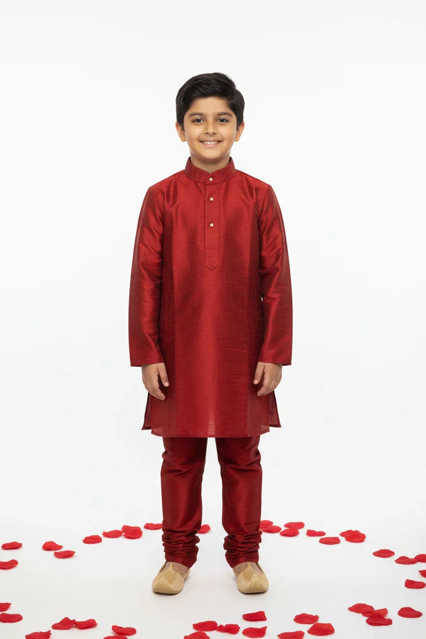 Smiling boy wearing red silk kurta pajama and beige traditional shoes, standing on white floor with red rose petals