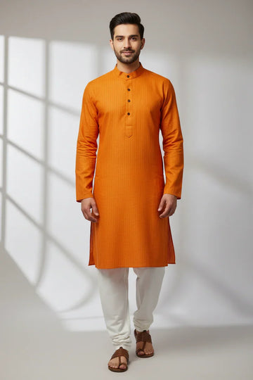 Man wearing orange Indian ethnic kurta and white churidar pants, standing in a studio with soft window light