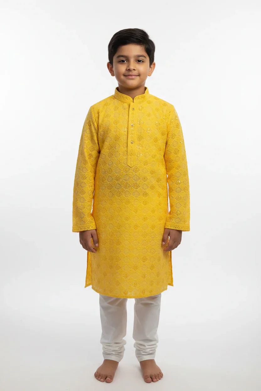 Young boy wearing yellow embroidered Indian kurta with white churidar pants standing barefoot on white background