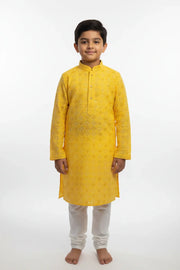 Young boy wearing yellow embroidered Indian kurta with white churidar pants standing barefoot on white background
