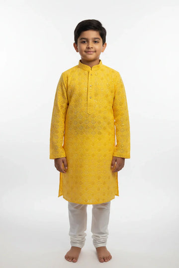 Young boy wearing yellow embroidered Indian kurta with white churidar pants standing barefoot on white background