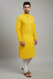 Man wearing yellow embroidered Indian kurta with white churidar pants and brown sandals against grey backdrop