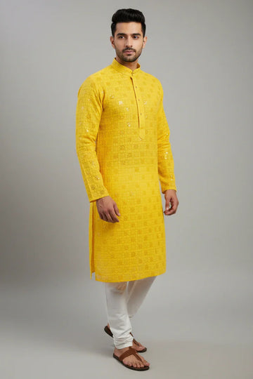 Man wearing yellow embroidered Indian kurta with white churidar pants and brown sandals against grey backdrop