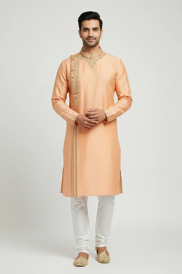 Man wearing peach embroidered Indian kurta with white churidar and traditional jutti shoes, studio background