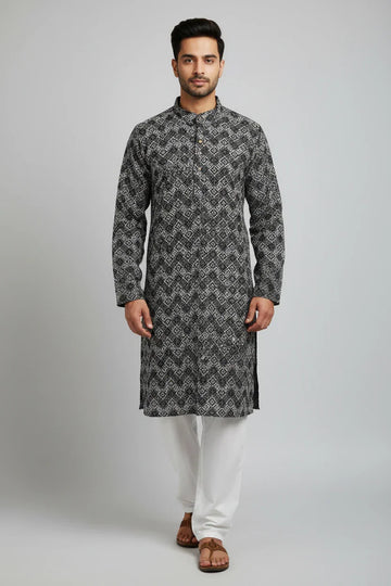 Man wearing black and white patterned Indian kurta with white pants and brown sandals on plain background