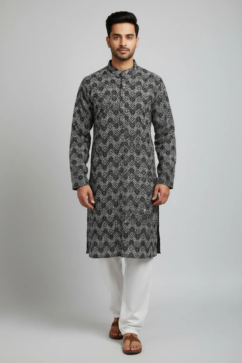 Man wearing black and white patterned Indian kurta with white pants and brown sandals on plain background