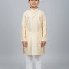 Young boy wearing cream and gold patterned Indian kurta with white churidar pants on gray background