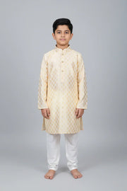 Young boy wearing cream and gold patterned Indian kurta with white churidar pants on gray background