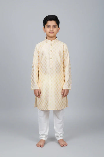 Young boy wearing cream and gold patterned Indian kurta with white churidar pants on gray background