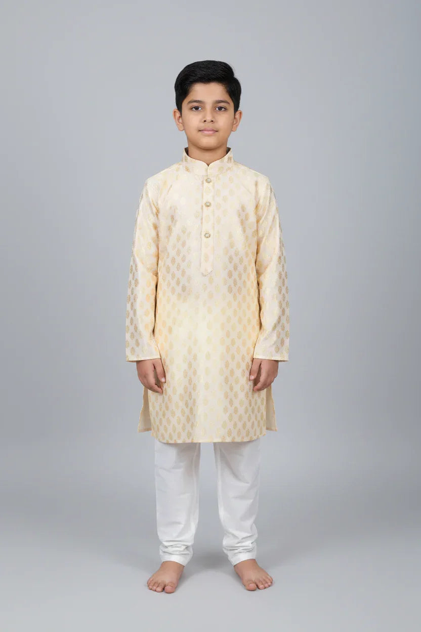 Young boy wearing cream and gold patterned Indian kurta with white churidar pants on gray background
