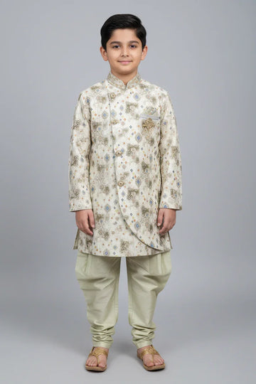 Boy wearing cream-colored traditional Indian sherwani with intricate patterns and churidar pants at Anandi Fashions